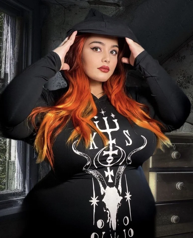 A woman with long red hair and dramatic makeup holds a black hat on her head. Text reads, Lilith Fury, Horror actress, disabled plus size model, and body positivity activist.