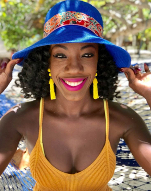 On a sun-dappled patio, a smiling woman grasps her vibrant blue hat. She wears hot pink lipstick, bright yellow earrings and knitted top.