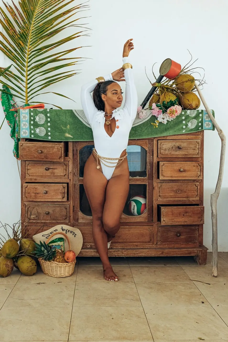 A woman in a long-sleeved, high-cut white one-piece swimsuit, accessorized with a gold chain belt, poses leaning against a rustic wooden cabinet. The scene is decorated with tropical elements like palm fronds and coconuts.