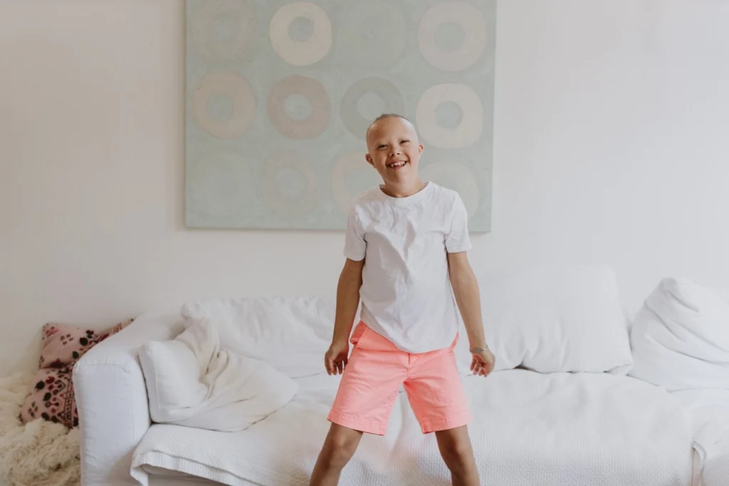 A joyful young boy with alopecia wearing a white t-shirt and bright coral-colored shorts. He is captured mid-motion, looking at the camera with a big smile while standing or jumping on a large white sectional sofa. Behind him, a light-colored abstract painting with circular patterns hangs on a plain white wall, creating a bright and airy indoor setting.