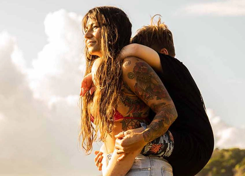 Against a cloudy sky, a woman with long hair and tattoos on her bare arms, carries a boy on her back. Niki & Jimmy: disability, love, laughter, happiness.