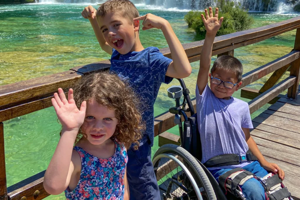 Three kids are on a wooden bridge overlooking clear green waters and a distant waterfall. The girl holds up a hand in a wave. A bigger boy smiles and holds both hands up in a muscle pose. A small boy raises one arm high as he sits smiling in his wheelchair. Text reads, Tips for traveling with spina bifida by Jennifer Allen. Read More.
