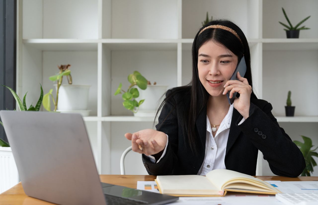 A smiling woman talks on her cell phone, her laptop open on the table in front of her, her hand open towards it. Behind her are white cubbies filled with potted plants.