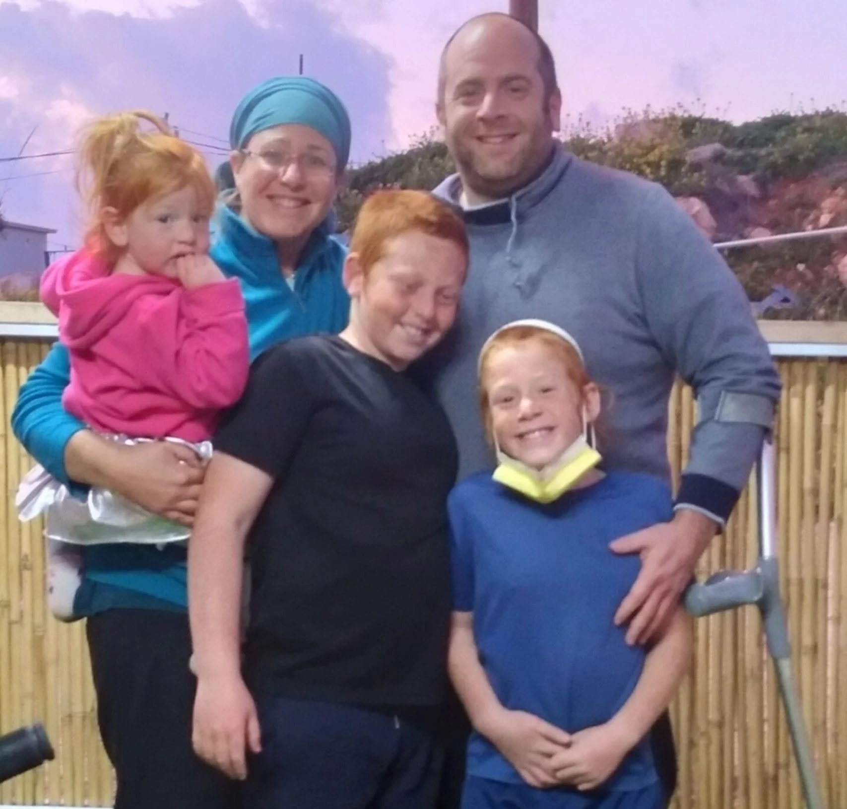 A family portrait showing two adults and three children posing outdoors in the evening. The mother, wearing a blue head covering and jacket, holds a small girl in a pink top. The father, wearing a gray sweatshirt, stands with his arm around a boy and a girl, both with bright red hair and wide smiles. A crutch or walking aid is visible by the father's right leg.