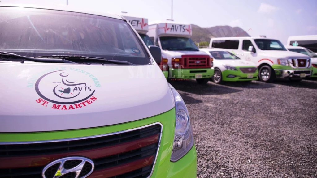 A line of airport or specialized transport vehicles, including vans and cars, parked in a gravel lot on a sunny day. The foreground focuses on the hood of a white and bright green Hyundai van, which features a circular logo for "Accessible Ventures AVTS St. Maarten."