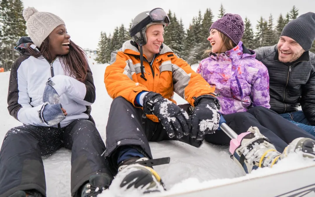 Three young people in ski gear sit in the snow, sharing a laugh.
