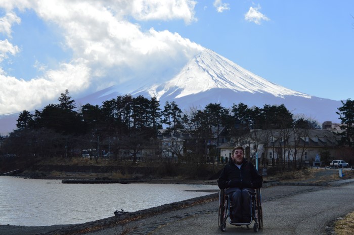 A man in a wheelchair sits on a path, with water on one side of him and a majestic snow-capped mountain in the distance.