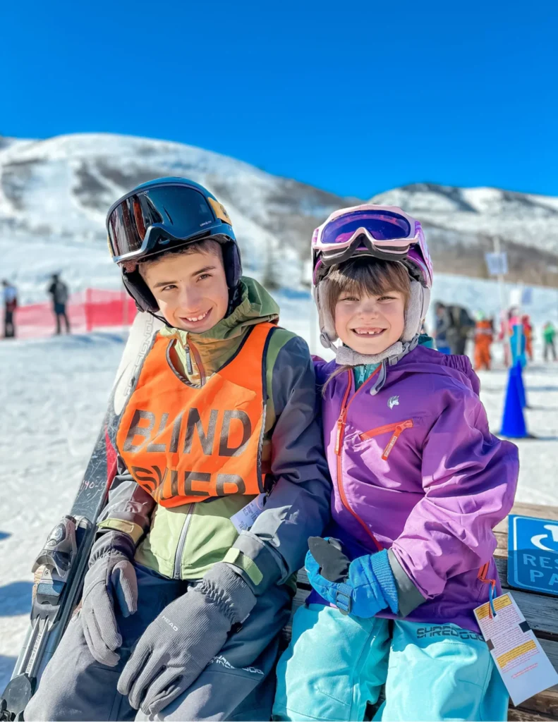 It's a sunny day on a ski mountain as two kids in ski gear smile at the camera.