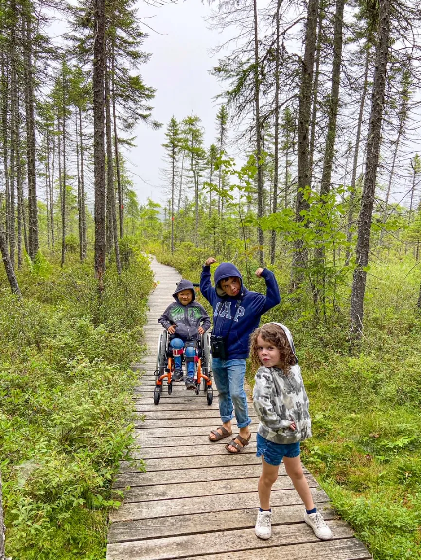 Three kids pose on a wooden walkway in a forest; two standing and one in a wheelchair.