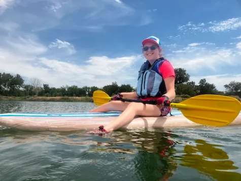 A sunny blue sky holds wisps of clouds. A girl sits smiling in a kayak, her feet in the water of a lake, a yellow paddle across her body.