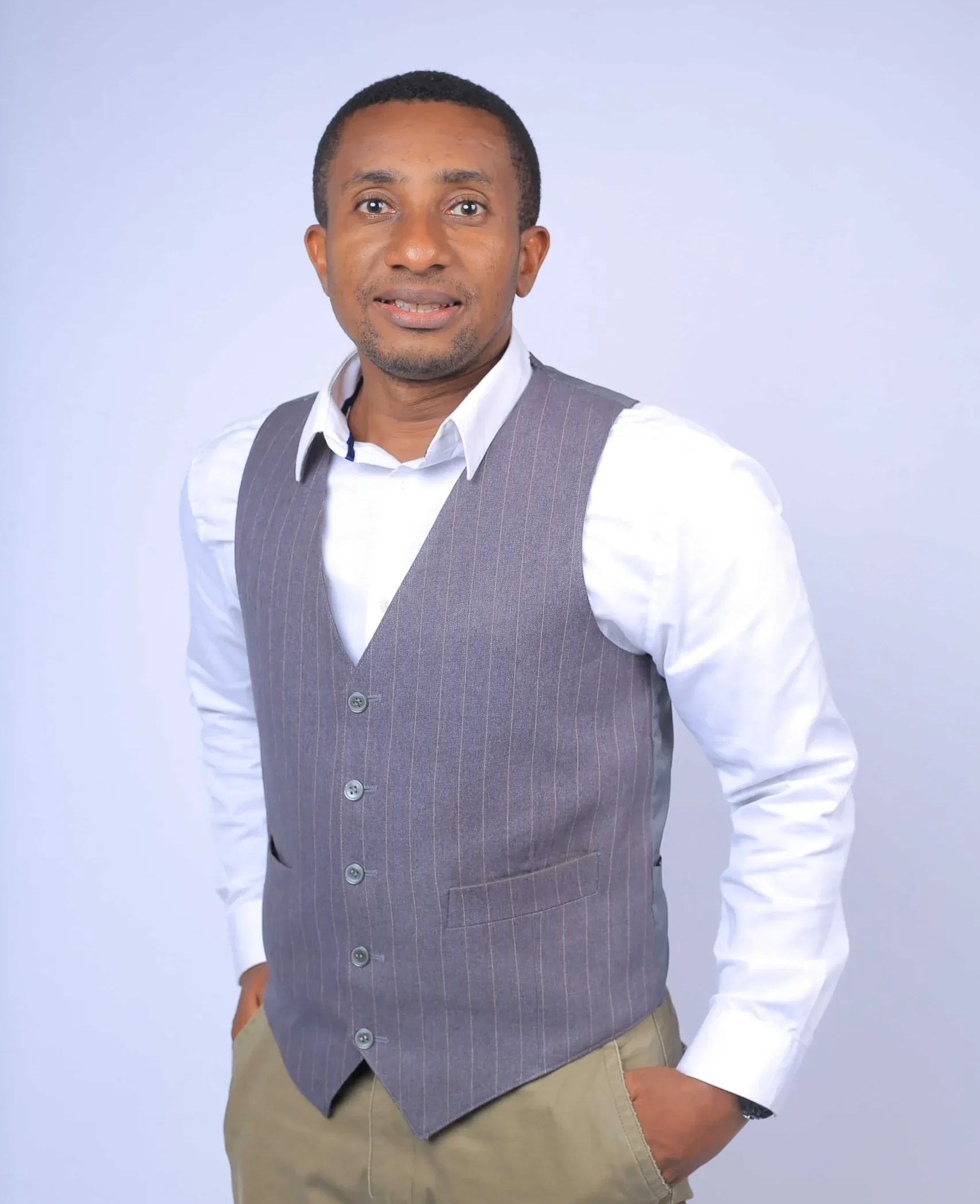 A professional headshot of a smiling man with a light brown complexion and short dark hair. He is wearing a white collared shirt, a grey pinstripe vest, and tan trousers. He stands with his hands in his pockets against a plain white or light grey background.