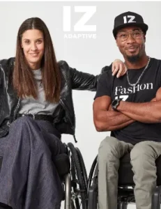 A promotional photo for IZ Adaptive clothing featuring two models sitting side-by-side in wheelchairs. On the left, a woman with long dark hair wears a gray top, dark wide-leg pants, and a black leather jacket. On the right, a man with a beard and dark glasses wears a black cap and a black t-shirt that says "Fashion IZ" and has his arm resting on the woman's shoulder. The text "IZ ADAPTIVE" is visible in the background above them.