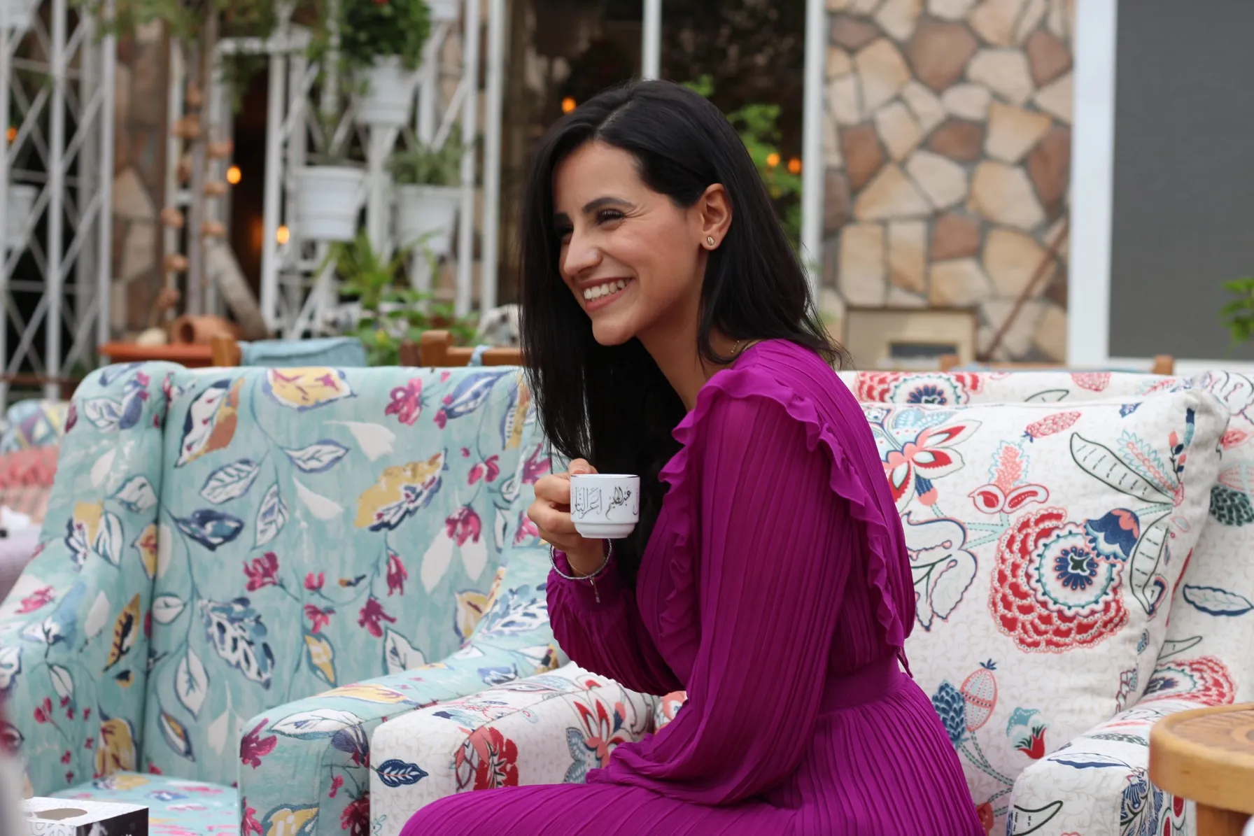 A beautiful young woman with long dark hair, wearing a vibrant, long-sleeved, pleated magenta dress with ruffled details, sits on a colorful floral print outdoor sofa. She is smiling broadly and looking off to the side while holding a small white coffee cup. The background shows a relaxed, stylish outdoor setting with stone-patterned walls and hanging lights.