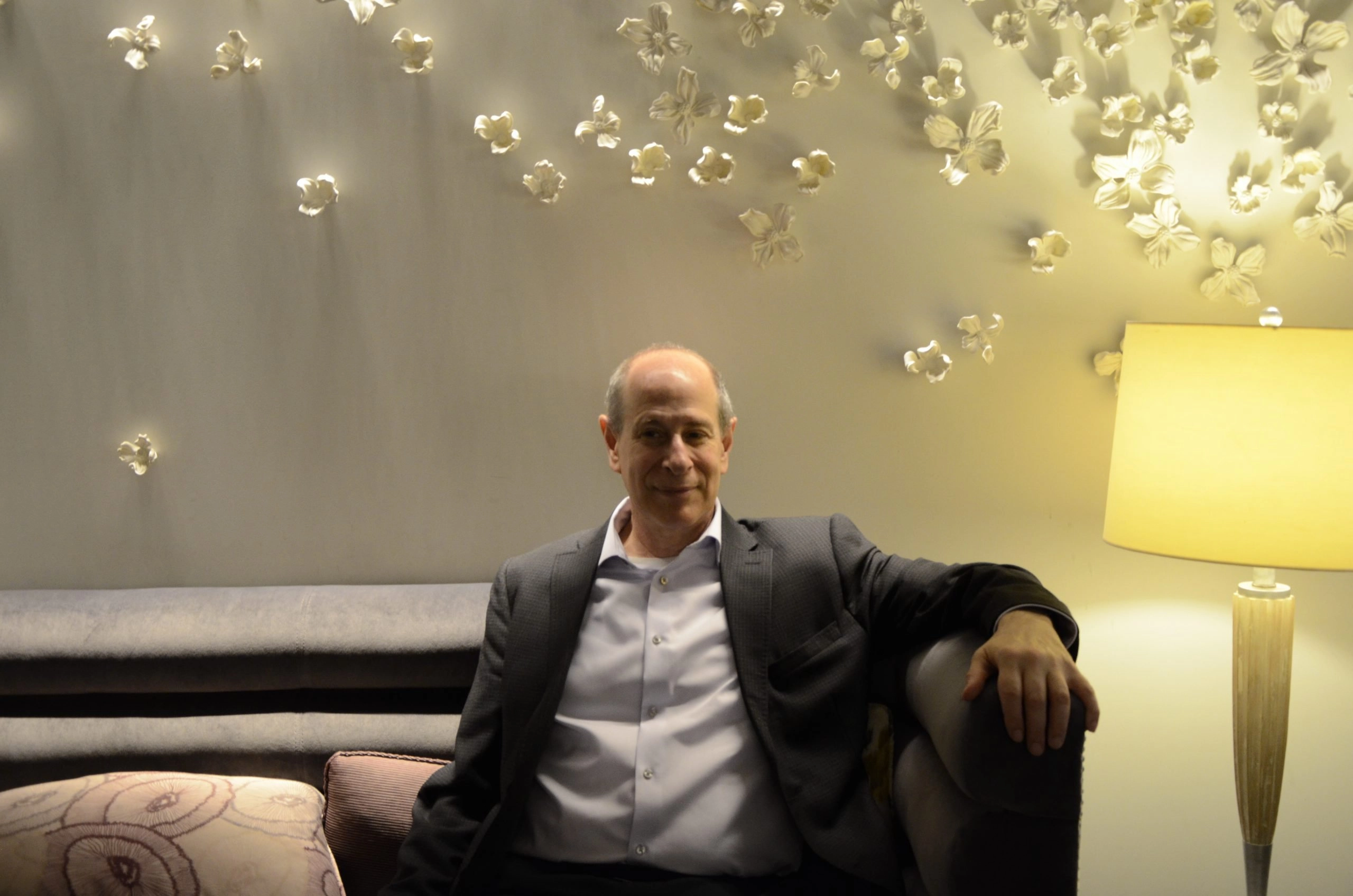 A middle-aged, balding man in a dark suit and white shirt sits on a dark sofa, leaning his arm on the backrest, and smiling slightly. He is illuminated by a yellow-toned lamp on the right and spotlighting on the white wall behind him, which is decorated with numerous white, three-dimensional flower sculptures.