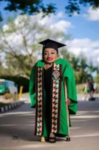 A woman with achondroplasia, dressed in a black graduation gown and a bright green academic hood featuring black, red, and geometric patterns, stands outdoors on a paved area. She is wearing a black mortarboard cap and is supported by a pair of crutches. The background shows trees, a blue sky with white clouds, and the blurred shape of a white bus and other people in the distance, suggesting a university campus setting.