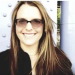 A headshot of a smiling woman with long brown hair wearing dark sunglasses and a black collared shirt, standing outdoors against a light-colored structure featuring large rivets.