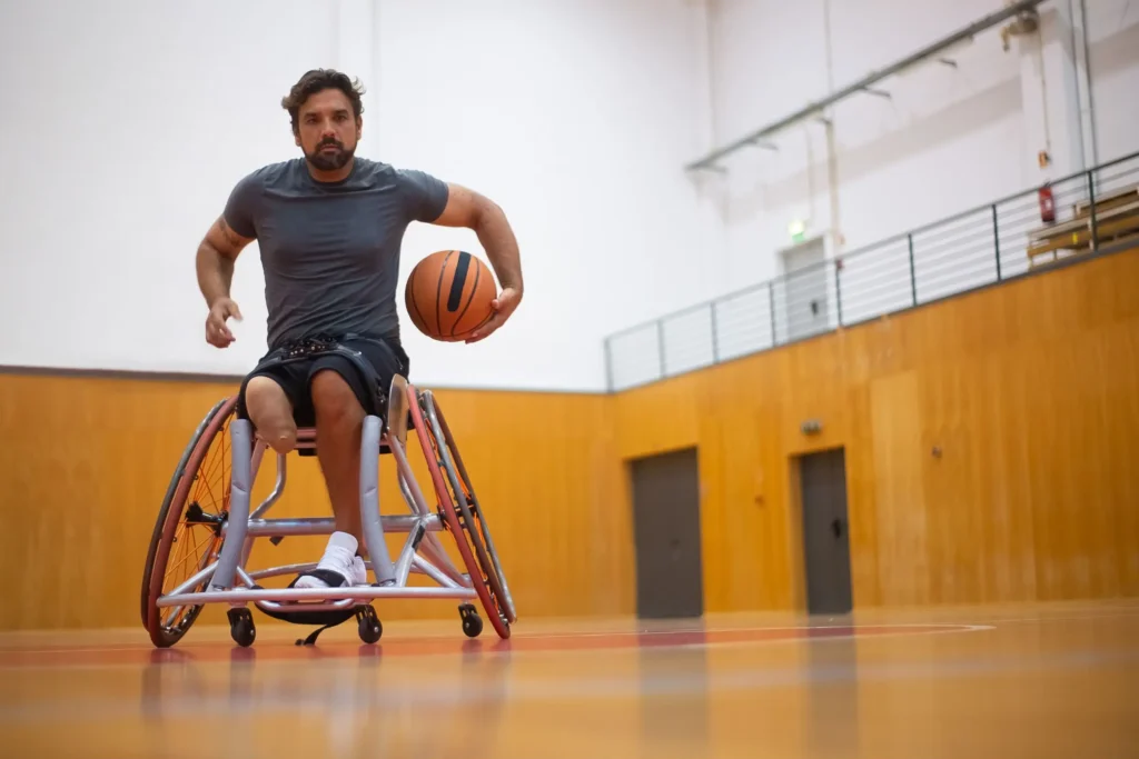 A man with one amputated lower leg sits in a sports wheelchair wielding a basketball on an indoor court.