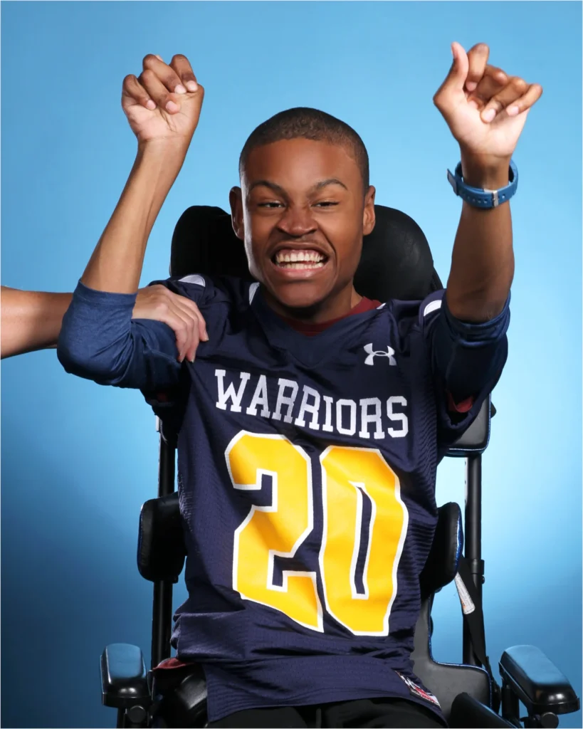 A young male athlete with a close-cropped haircut, seated in a power wheelchair, wearing a navy blue football jersey with the word "WARRIORS" and the number "20" in yellow. He is cheering with his teeth clenched and both hands raised in the air, with another person's hand supporting his left arm. The background is a solid bright blue.