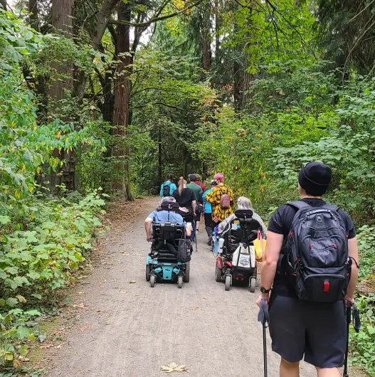A group of people hike over a forest path. Some walk, some use walking sticks, some are in wheelchairs.
