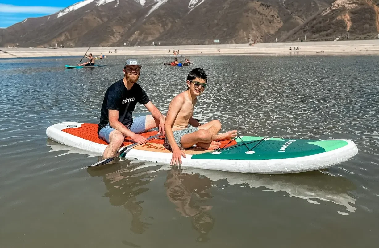 Two people sitting on a paddleboard on a lake with snow-capped mountains and a dam wall in the background.