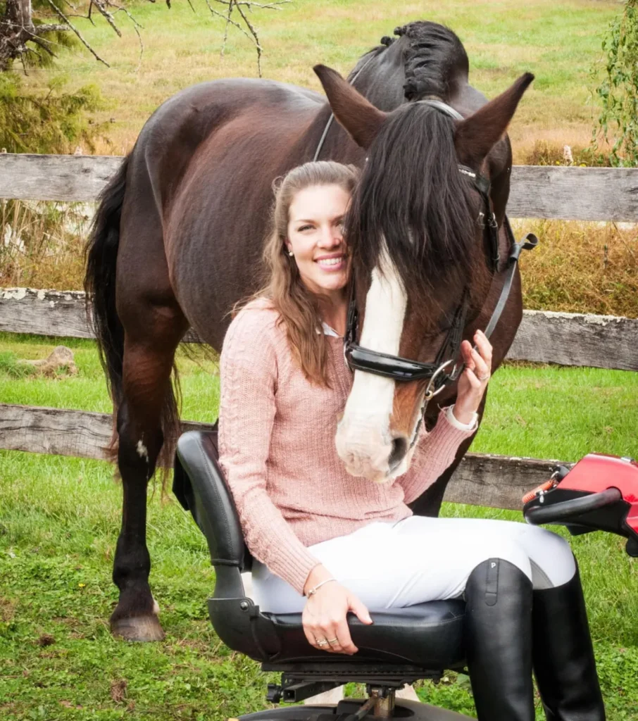 A smiling woman with long brown hair sits on a black-seated mobility device in a grassy outdoor area. She is wearing a light pink cable-knit sweater, white riding pants, and tall black riding boots. She is embracing the head of a tall, dark brown horse with a white blaze, who is nuzzling her cheek. A rustic wooden fence is visible behind them.