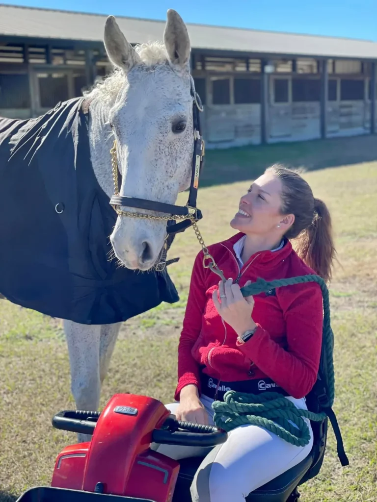 A smiling woman with her hair pulled back sits on a small red mobility scooter, looking affectionately up at a tall white horse. The horse wears a black turnout blanket and a bridle with a gold chain. The woman is wearing a red jacket and white riding pants, holding a green rope. They are outdoors on a grassy field in front of a large barn structure under a bright, sunny sky.
