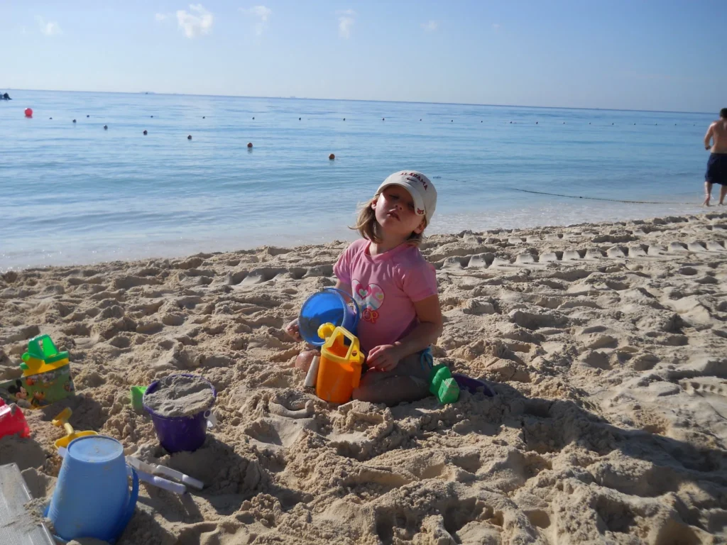 A young girl with blonde hair, wearing a pink t-shirt and a light-colored baseball cap, sits on a sandy beach looking up toward the sky with a slight squint. She is surrounded by several colorful sand pails and shovels. The beautiful blue ocean stretches out behind her under a clear sky, with several buoys visible in the water and a person standing in the far right background.