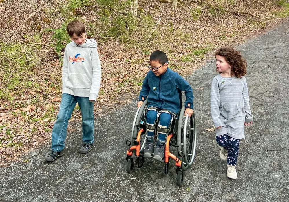 Three children walk on a paved trail through a bare, early spring forest. A boy on the left wears a grey hoodie and jeans. In the center, a boy with glasses is seated in a lightweight manual wheelchair with orange accents, wearing a dark blue jacket and leg braces. A girl with curly hair walks on the right, holding a small stick. The woods rise steeply behind them with tall, leafless trees.