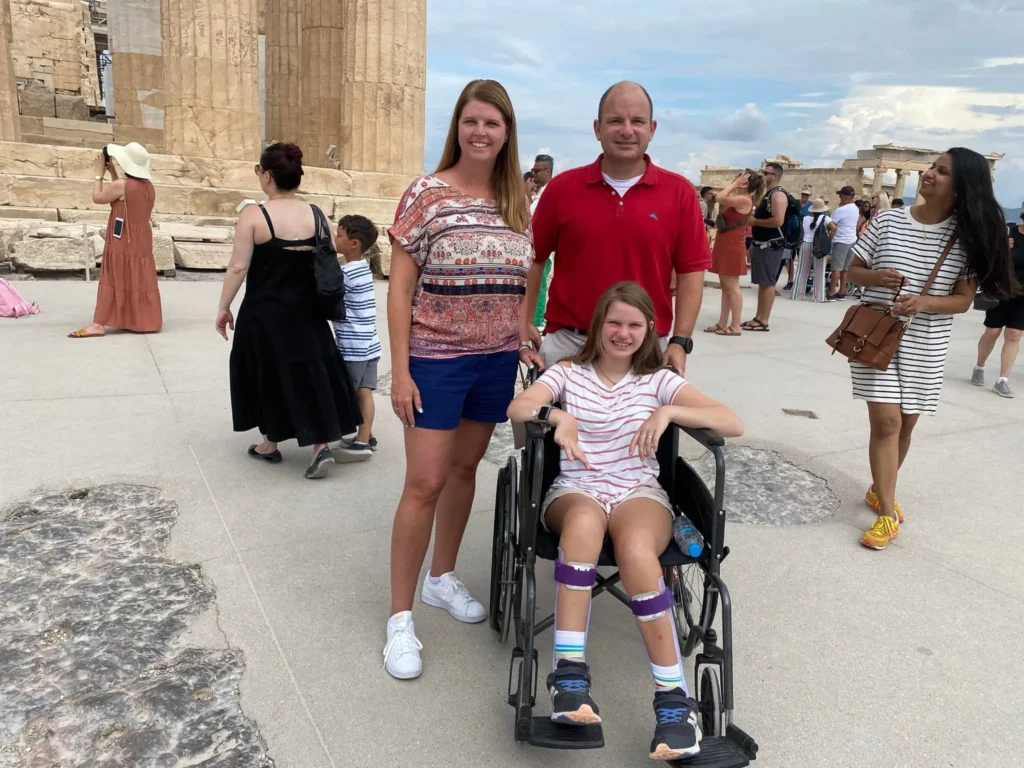A family of three poses for a photo at a crowded historical site, likely the Acropolis in Athens, Greece, with ancient stone columns visible behind them. The father wears a red polo shirt, and the mother wears a patterned top and shorts. Their daughter is seated in a manual wheelchair, smiling, and wearing a striped t-shirt and shorts with braces or supports on her lower legs and ankles. Tourists are visible all around them in the background.