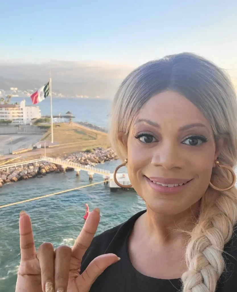 A selfie of a smiling woman with a blonde braid and gold hoop earrings, wearing a black top. She is holding her hand up in the "I love you" sign (or "rock on" sign). She is standing on a cruise ship or port with the ocean and a coastal town in the background. A Mexican flag is visible flying over the port.