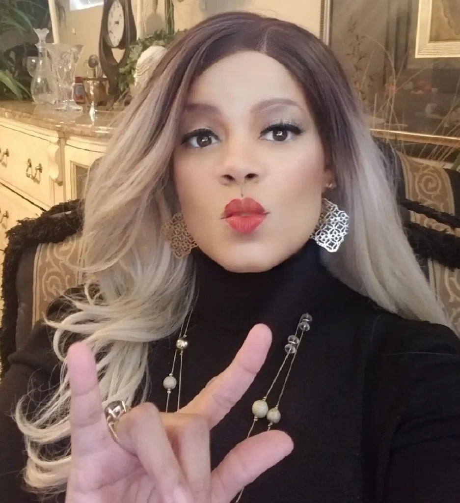A woman with long, brown-to-blonde ombré hair, wearing a black turtleneck sweater, poses indoors. She has red lipstick on and is making a kissing face while holding her right hand up in the "I love you" sign (or "rock on" sign). She is wearing large, decorative silver earrings and long, beaded gold necklaces. In the blurry background, there is an ornate, cream-colored cabinet and a clock.