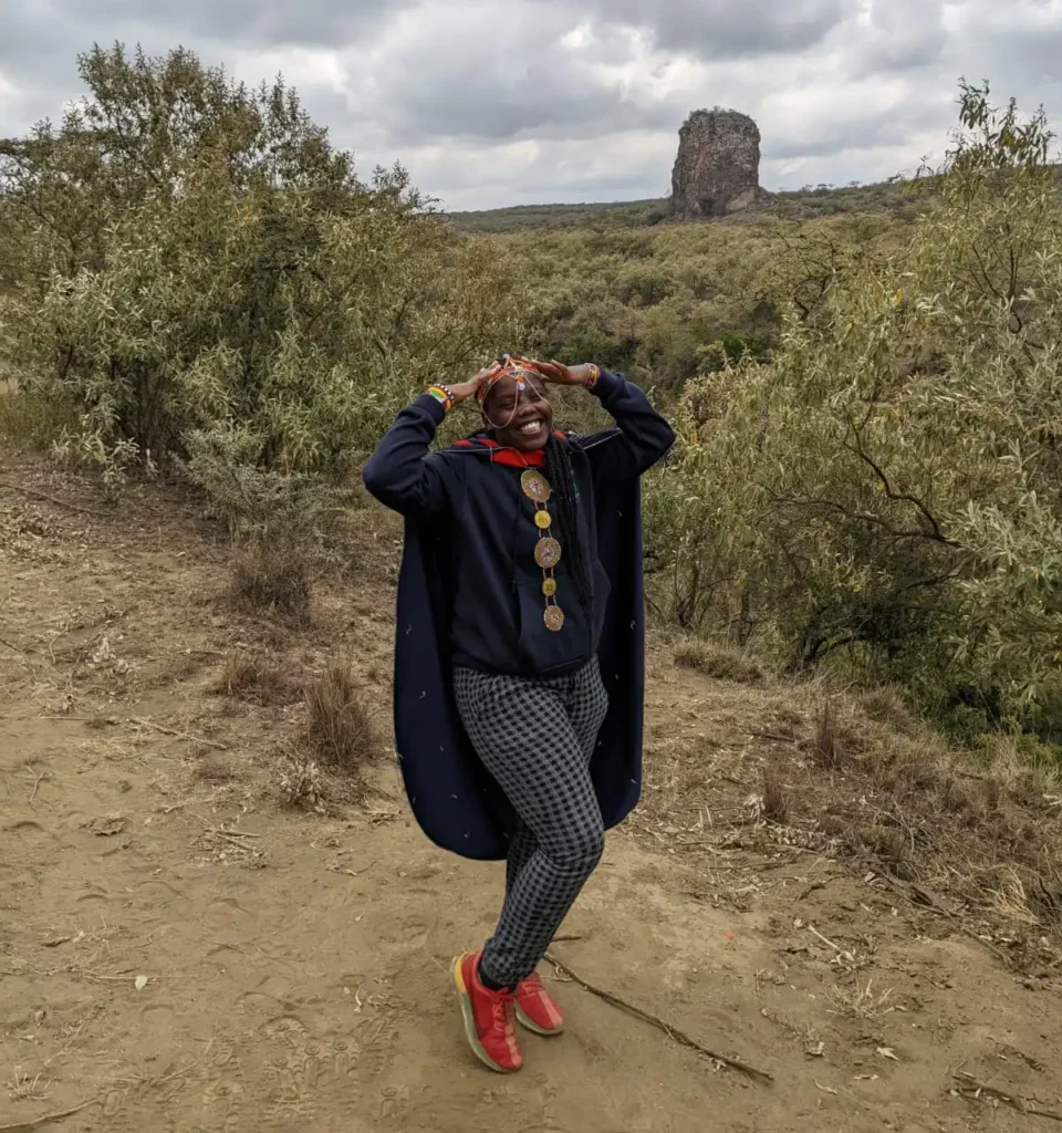 A person with facial paint, wearing a dark blue hoodie, a long navy cape, checkered grey pants, and bright red shoes, stands on a dry dirt path outdoors. They are smiling and holding their hands up to their head. In the background, there is dense, dry shrubbery and a distinctive, large, rocky spire or formation rising up from the landscape under a cloudy sky. The setting appears to be a dry, bushland environment, possibly in Kenya near a feature like the Pride Rock inspiration.