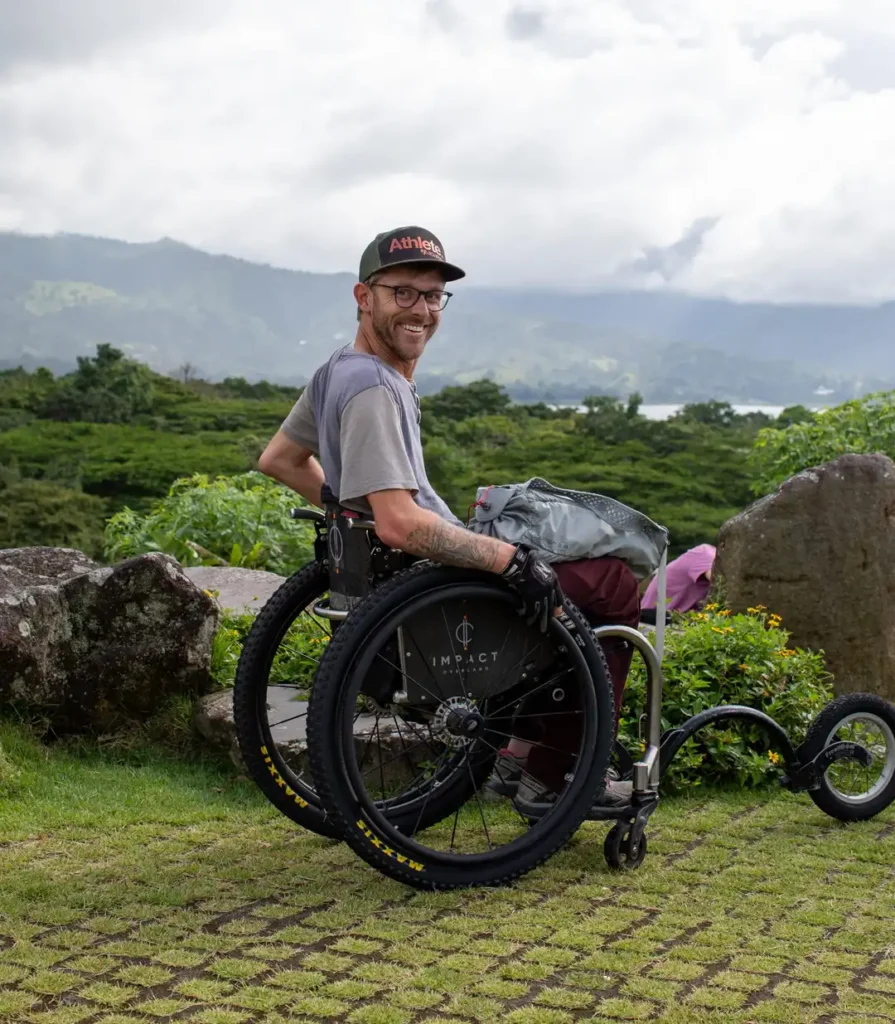 A smiling man with a beard and glasses, wearing a baseball cap and a gray t-shirt, is seated in an all-terrain wheelchair outdoors. The chair has large, knobby tires and a small front wheel attachment. The background is a lush, green, hilly landscape under an overcast sky, with a body of water visible in the distance.