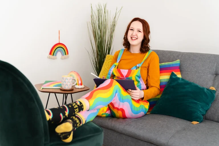 A woman sitting on a grey sofa with colorful decor. She has curly auburn hair, wears a mustard yellow long-sleeve shirt and vibrant multicolored overalls. She is holding a digital tablet with a stylus, looking away from the camera with a joyful smile. Around her are rainbow-themed items—a rainbow wall hanging, a rainbow-striped notebook, and a rainbow-colored cup. Her socks are black with colorful accents, complementing the overall cheerful and bright theme of the setting.