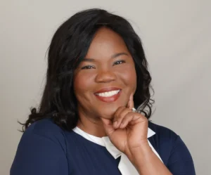 The image shows a smiling woman with medium-length dark hair. She's wearing a navy blue dress with white cuffs and a white collar, which gives her a professional and put-together appearance. The woman is resting her face on her hand, her elbow bent and fingers pointed upward. Her pose exudes confidence and friendliness, and she appears to be in a professional setting, suggesting she might be in a role that involves engagement with people, such as a consultant, educator, or business leader. The lighting is soft and flattering, highlighting her features without creating harsh shadows.