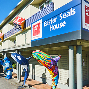 A vibrant photograph of Easter Seals House entrance in Vancouver, BC. To the left, a distinctive blue sign with white lettering reads "Easter Seals House" above a smaller sign for "Lions" with its logo. In front of the building, three whimsical, colorfully painted orca whale statues in various poses add a playful and artistic touch to the scene. The clear sky suggests a bright, sunny day.