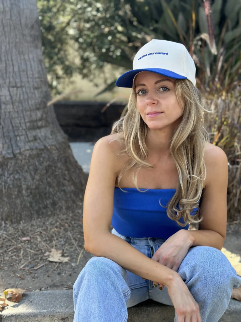 A woman sitting outdoors with a casual pose. She's wearing a blue tank top, light-wash denim jeans, and a white and blue baseball cap with the text "caption your content" on it. Her blonde hair is styled in loose waves, and she looks contemplative or relaxed. In the background, there's a tree and some shrubbery, indicating a park or a similar green space.
