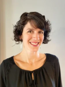 A woman with a cheerful expression, sporting short curly hair and wearing a black top with a round neckline and a small keyhole detail. She is accessorized with dangle earrings and the background is neutral, providing a professional setting. Her eyes are bright, and she is offering a friendly smile to the viewer.