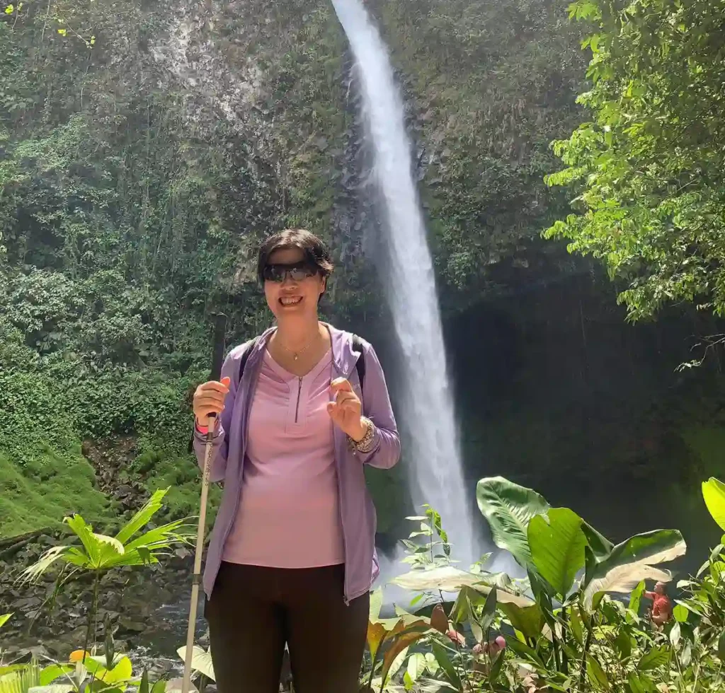 A blind lady stands smiling in the foreground, wearing sunglasses, a light purple top with a darker purple zip-up jacket, and dark pants. They hold a walking stick in one hand and give a thumbs-up with the other. Behind them, a majestic waterfall cascades down a rocky cliff amidst lush green vegetation. Sunlight filters through the tree leaves above, adding a bright, natural glow to the serene landscape.