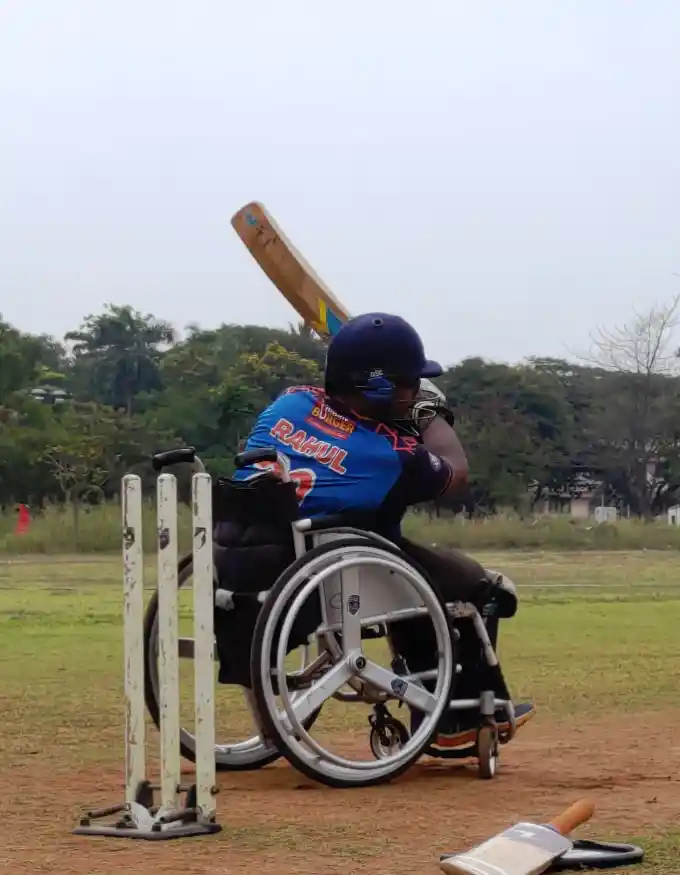 This image shows a wheelchair cricket player on the field. The player is donned in a blue and orange cricket jersey with the name "RAHUL" printed on the back, indicating either the player's name or a sponsored athlete. The player is wearing protective gear, including a helmet and pads, which suggests they are batting. The cricket bat is captured mid-swing, implying that the player has just hit the ball. Three stumps with bails are present, which represent the wicket in cricket. Another bat lies on the ground, suggesting that the player might have changed bats or there was another player present earlier. The background is an open field with trees and a hazy sky, typical of an outdoor cricket field.
