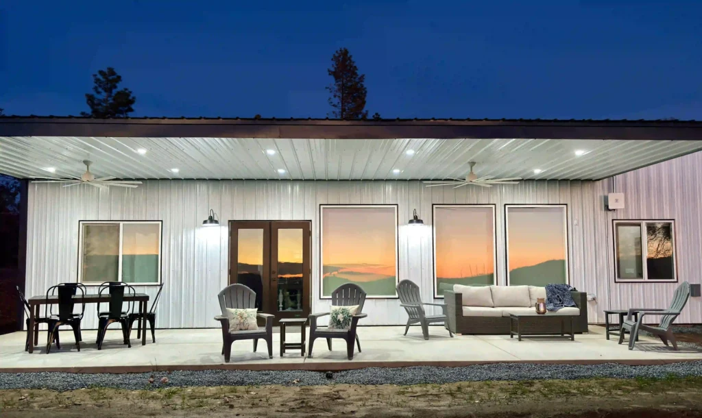 The image shows a well-lit back patio of a modern house at sunset. The house exterior features a metal facade with a covered patio area. There are several seating arrangements including a dining table with metal chairs, Adirondack chairs, and a cushioned outdoor sofa with a coffee table. The patio is adorned with pillows and blankets, giving it a cozy ambiance. Ceiling fans are installed above, and wall-mounted lights are illuminated, casting a warm glow. The large windows and glass doors reflect the vibrant sunset sky, adding to the serene and picturesque setting.