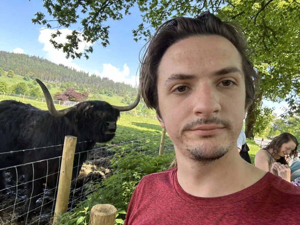 A man in a red shirt is taking a selfie outdoors. Behind him, a large black Highland cow with long horns is standing in a fenced grassy area. The background shows a lush green landscape with trees, hills, and a clear blue sky. To the right of the man, a woman in a white shirt is standing, possibly taking a photo or looking at something.