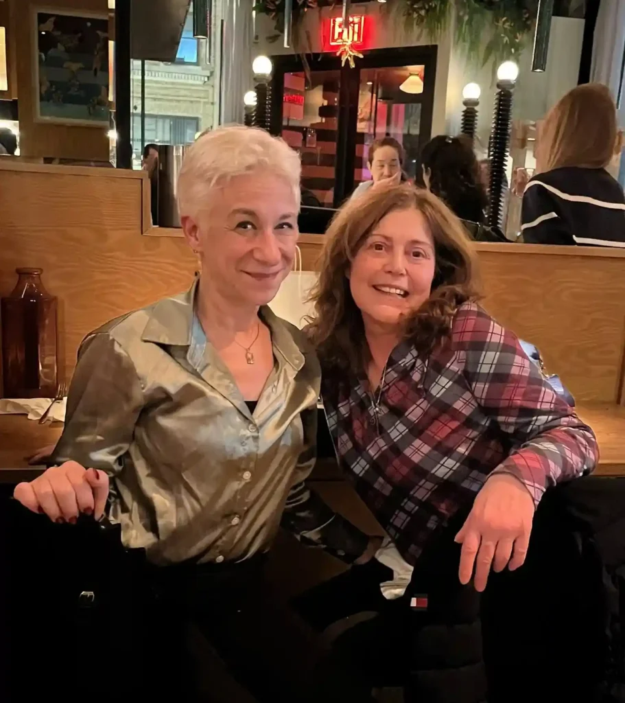 A photograph of two women sitting closely together in a cozy, dimly lit restaurant. The woman on the left has short, silver hair and is wearing a shiny, metallic blouse with a slight smile. The woman on the right has medium-length brown hair and is wearing a red and blue plaid shirt, smiling broadly. The background shows a mirror reflecting part of the restaurant interior, including hanging lights and other patrons. The atmosphere appears warm and friendly, with an inviting and relaxed setting.
