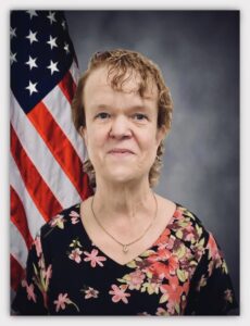 A professional portrait of a woman with short, curly hair, wearing a black floral blouse with pink and red flowers. She is standing in front of an American flag, which is partially visible on the left side of the image. The background is a gradient gray, giving the photo a formal and official appearance. The woman is smiling gently and wearing a small, elegant necklace.