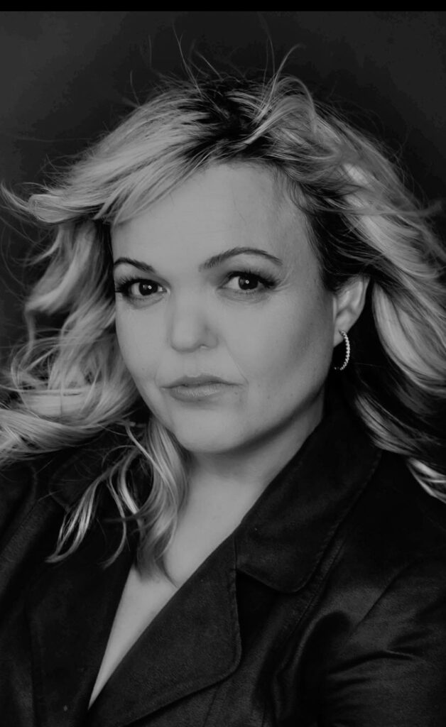 A black-and-white headshot of a woman with blonde, wavy hair. She has a serious expression, looking directly at the camera. She is wearing hoop earrings and a dark jacket. The photo has a high contrast with a sharp outline around her hair and upper body.