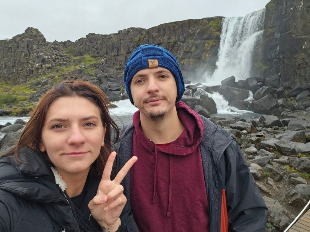A photo of two people posing in front of a waterfall. The person on the left is a woman with dark hair, making a peace sign with her hand. She is wearing a black jacket. The person on the right is a man wearing a blue beanie and a maroon hoodie with a black jacket over it. The waterfall and rocky landscape provide a scenic background.