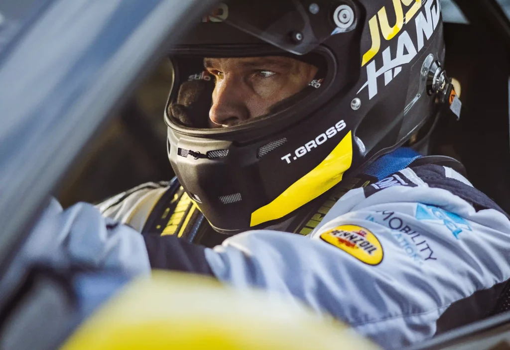 The image shows a race car driver seated in a car, intensely focused, wearing a black helmet with "T. GROSS" printed on the side in white text. The helmet features yellow accents and branding from various sponsors, including "Just Hands" at the top. The driver's suit also has sponsor patches, including a visible Pennzoil patch on the shoulder. The scene captures the driver's concentration, likely moments before or during a race. The lighting emphasizes the details on the driver's face, adding to the intensity of the moment.