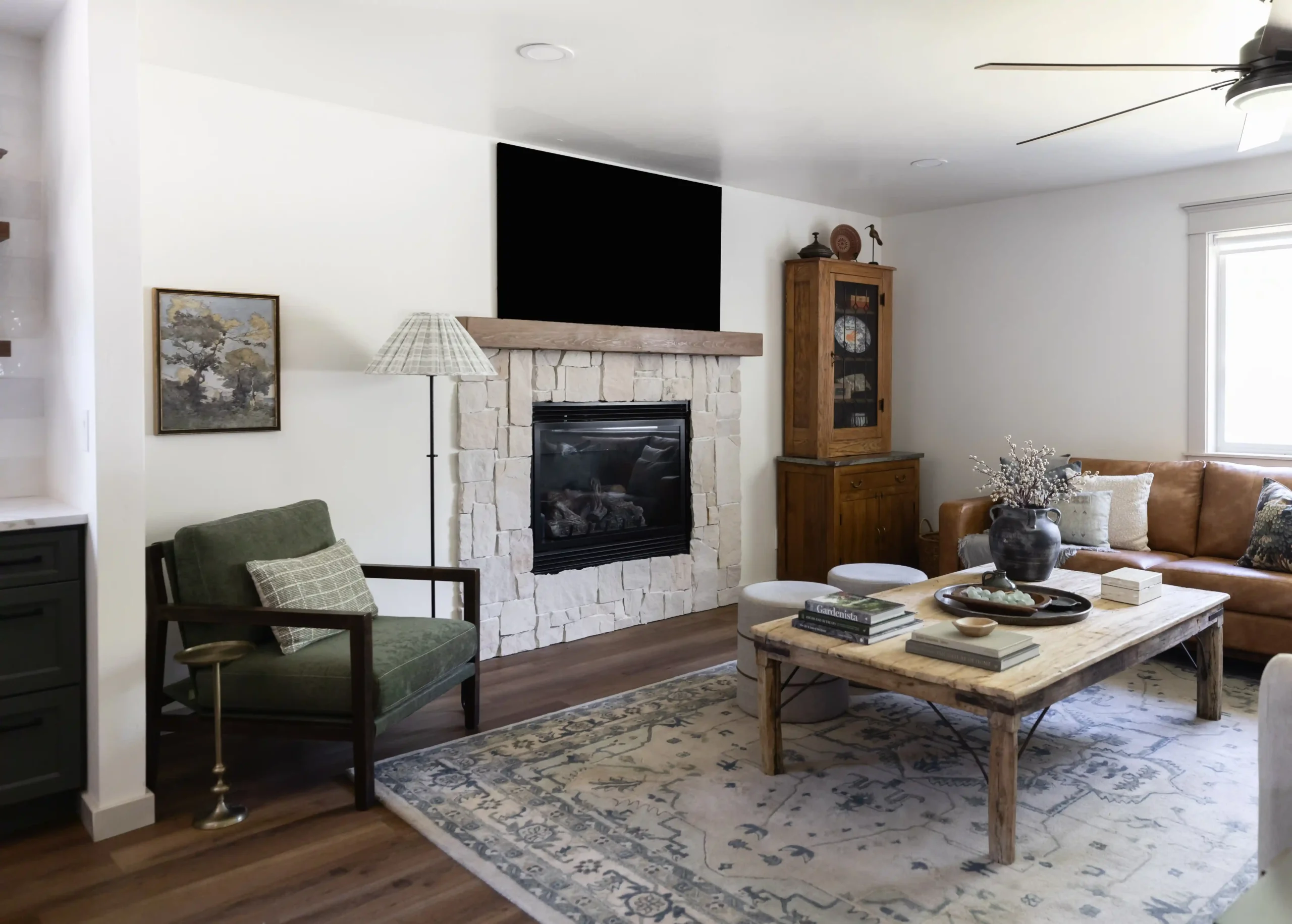 A cozy, rustic living room with a natural stone fireplace, topped by a wooden mantel and a black-framed TV. The space features a green armchair with a plaid pillow, a floor lamp with a patterned shade, and a leather sofa on the right. In the center, there is a large, wooden coffee table decorated with books, a vase with dried branches, and a ceramic bowl. A soft, patterned rug covers the hardwood floor. A wooden display cabinet stands next to the fireplace, adding to the room's warm and inviting ambiance.