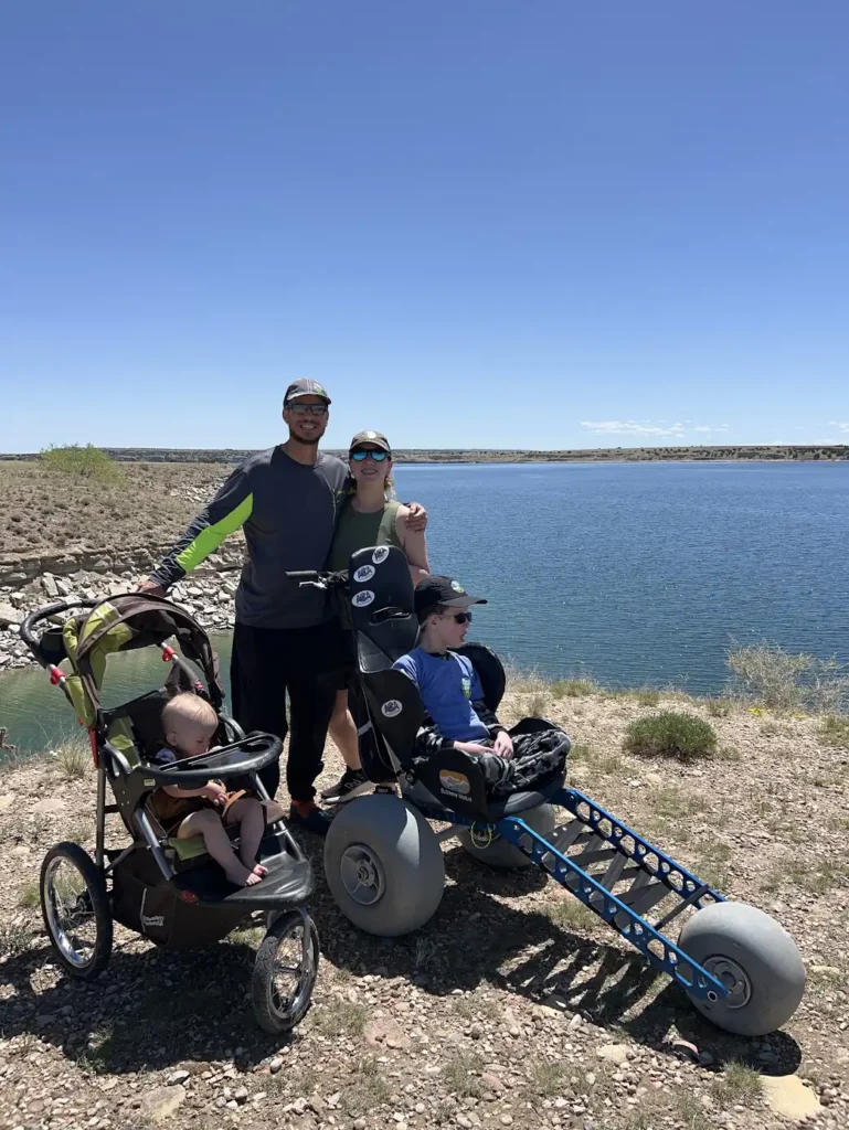 A family is outdoors near a lake on a clear, sunny day. Two adults are standing, one man and one woman, both wearing sunglasses and caps. The man has his arm around the woman. In front of them are two children in special strollers: one baby in a regular stroller on the left, and an older child in a larger off-road wheelchair with big balloon-like tires on the right. The scene shows a sense of adventure and togetherness, with a backdrop of blue water and a rocky shoreline.