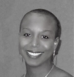 The image shows a black and white portrait of a smiling woman with short hair. She is wearing earrings and a necklace with large beads or ornaments. Her smile is warm, and she has an overall calm and pleasant expression.